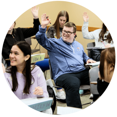 Image of a male student smilng with his hand raised with other students in th background in a classroom.